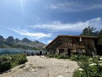 Vue de côté du refuge en bois, terrasse avec tables et chaises. Lac d'Allos visible à proximité, avec les Tours du lac en fond de photographie