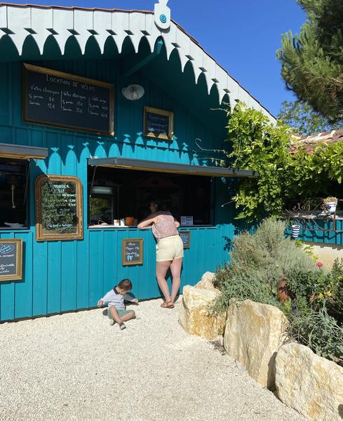 Une femme prenant une commande au bar, un grand chalet peint en bleu. Un jeune enfant est assis par terre.