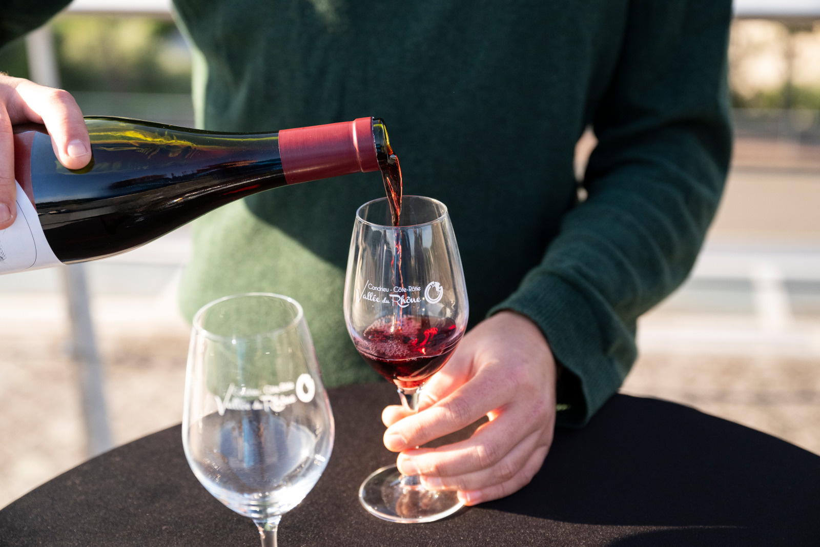 Deux verres de vin sont posés sur un table avec une nappe noire. Une personne est en train de verser du vin rouge dans l'un de ces verres