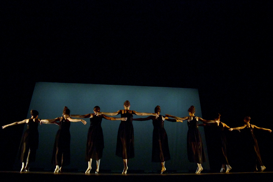 Photo prise lors du gala annuel de l'école de danse de Saint-Jean-en-Royans. 8 jeunes filles vêtues de longues robes noires se tiennent sur la pointe des pieds, toutes s'attrapent et regardent vers le sol. Elles sont de dos sur une scène éclairée de manière à ce qu'on ne voit qu'elles.
