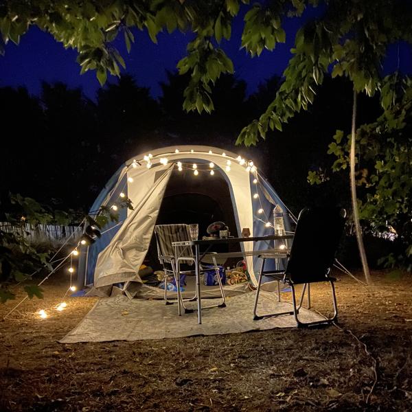 Une tente avec une guirlande accrochée sur l'entrée, la nuit. Une table et deux chaises de campings sont installés devant.
