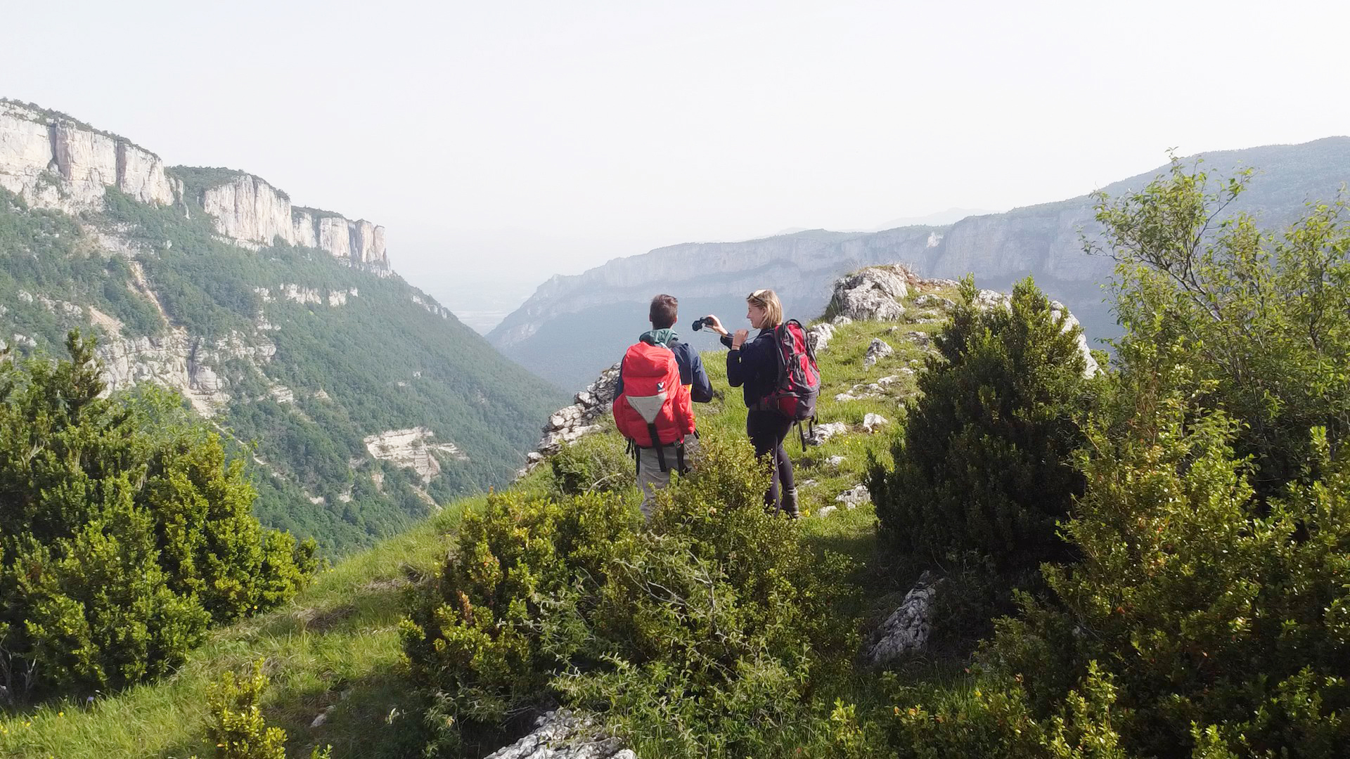 Deux randonneurs, sacs à dos sur les épaules, observent l’horizon à l’aide de jumelles depuis un promontoire, entourés par les montagnes