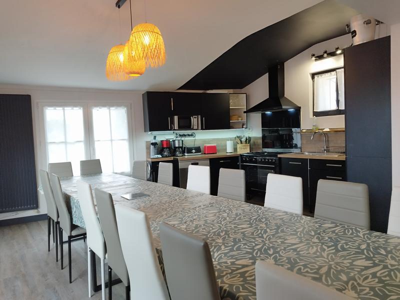 Une grande table avec une quinzaine de chaises, cuisine équipée ouverte.