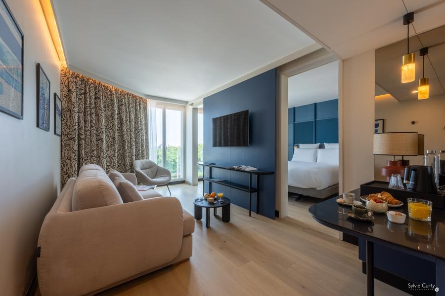 Une suite avec un chambre avec lit double séparée, un canapé, un fauteuil, une télévision, une table avec viennoiseries et boissons chaudes.