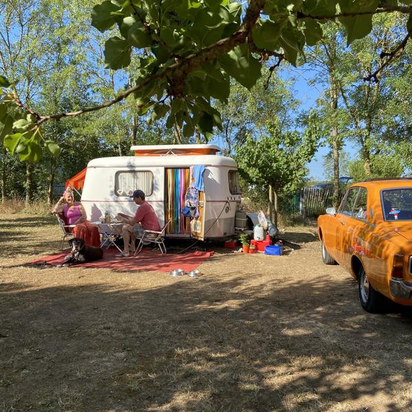 Un couple assis devant leur roulotte, une vieille voiture vintage à droite.