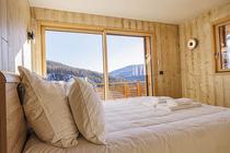 Chambre à coucher lumineuse lit deux places, ameublement bois, porte-fenêtre donnant sur un balcon en bois