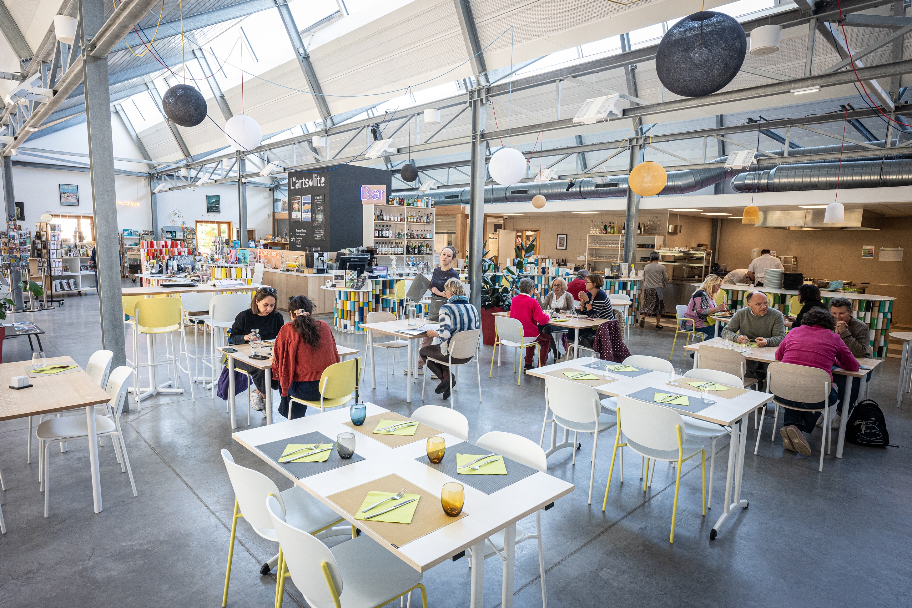 Des convives sont installés dans la salle de restauration avec en arrière plan les espaces cuisine, bar et boutique