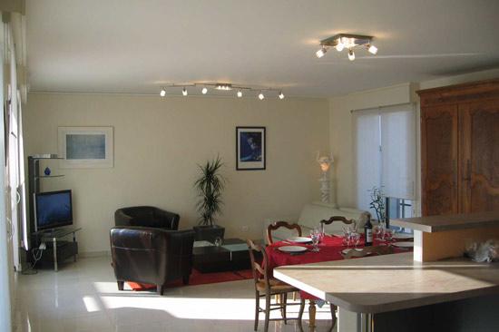 Autre vue de la salle à manger, avec une table dressée pour 6 personnes, et le coin salon en fond avec télévision, un canapé et deux fauteuils. Une grande armoire de famille en fond.