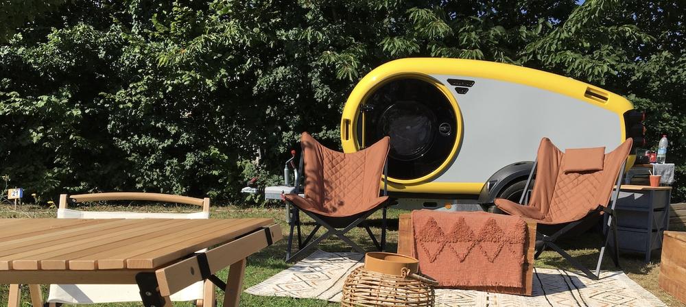 Une roulotte type futuriste derrière un coin cocooning avec des tables, fauteuils, tapis au sol.