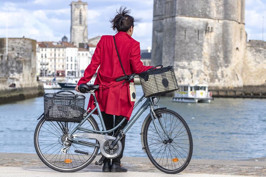 Une dame en rouge de dos, face au port, avec un vélo ayant des  paniers en matière de poches ostéricole à l'arrière et sur le guidon.