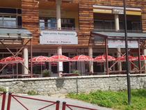Devanture du restaurant en été avec terrasse extérieure avec tables, chaises et parasols rouges. Panneau avec inscription : "Pourquoi pas ? Crêperie Pizza" "Spécialités montagnardes"