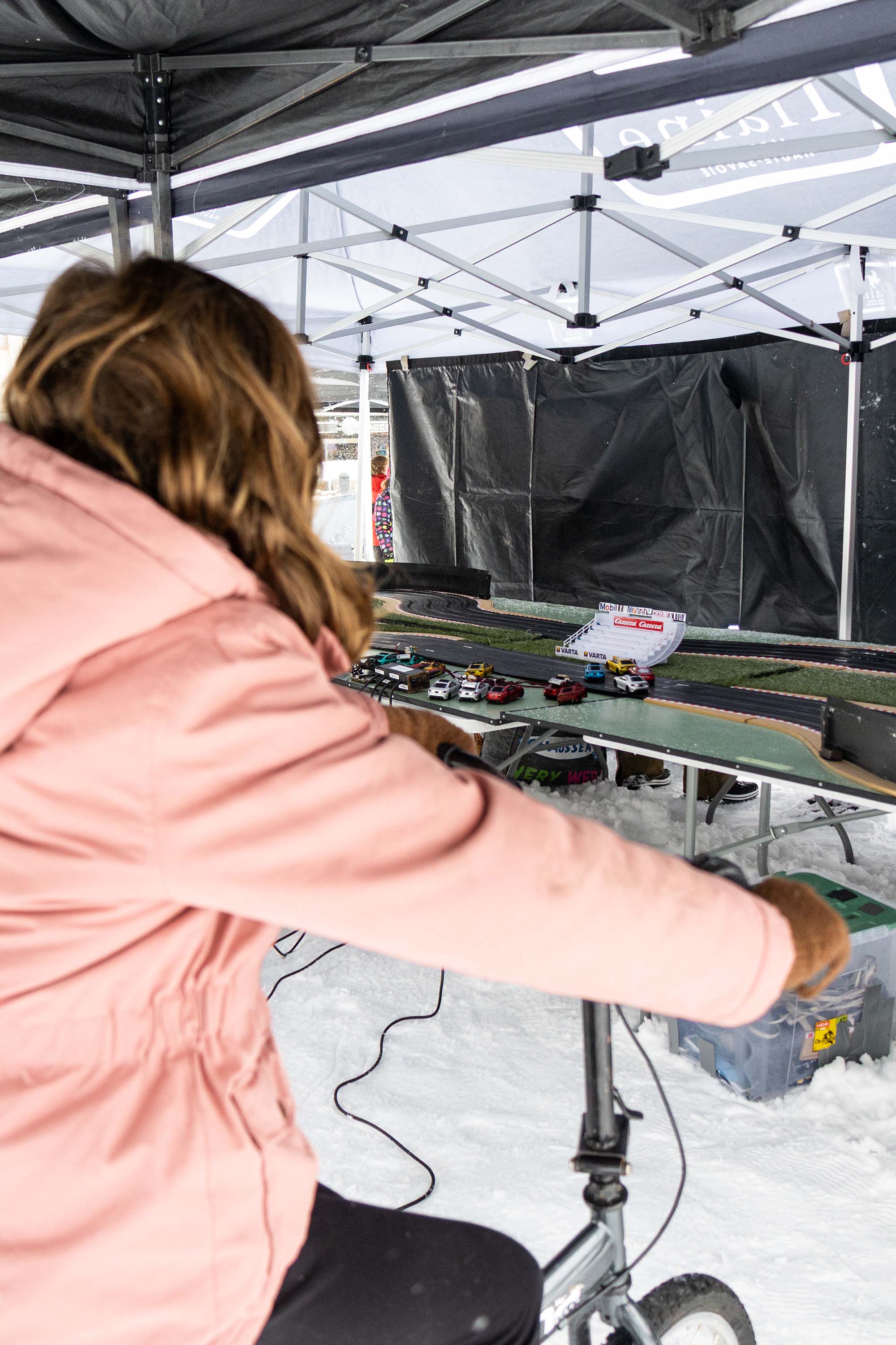 Une personne en veste rose pédale sur un vélo pour faire avancer des petites voitures électriques sur un circuit miniature.