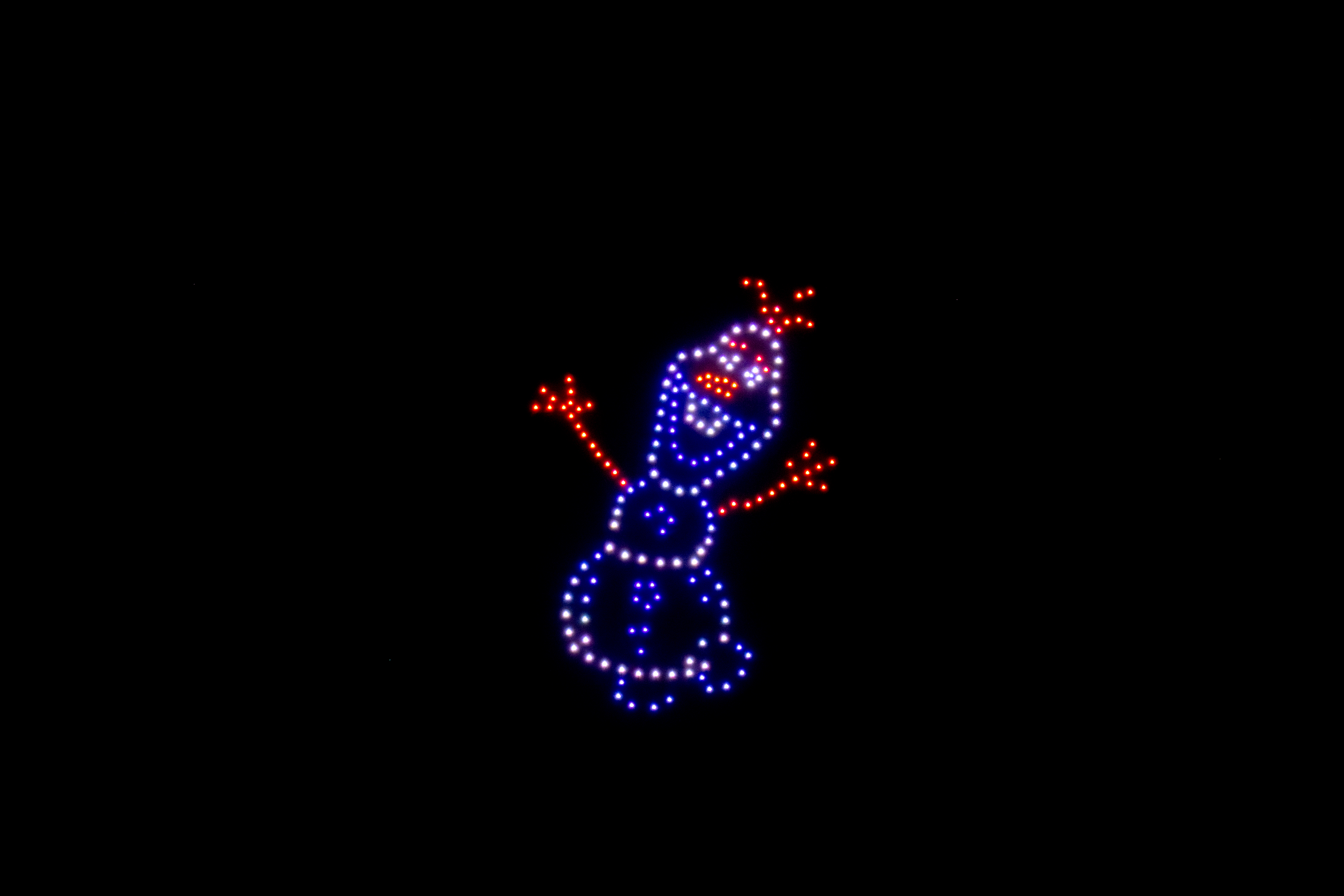 Spectacle de drones nocturne sur fond noir. Les points lumineux bleus, blancs et rouges dessinent le personnage d'Olaf, le bonhomme de neige de La Reine des Neiges, avec les bras levés.