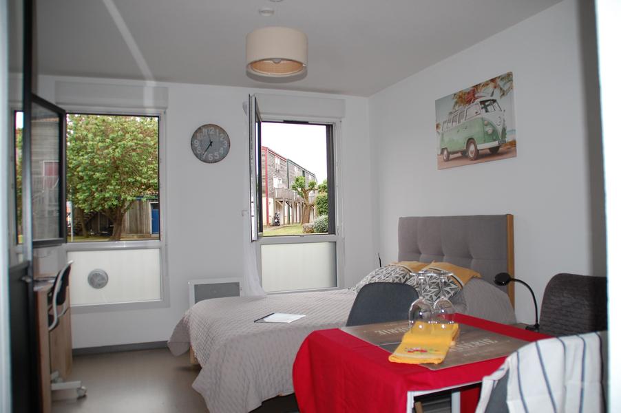 La pièce de vie avec lit double, table à manger, deux fenêtres ouvertes sur la rue.