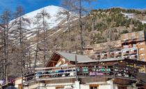 Vue d'ensemble du restaurant en bois, situé devant des immeubles de la station de ski, en haut d'un bâtiment sur deux niveaux, terrasse. Panneau "Sympa Snack" sur la devanture. Montagnes en arrière-plan