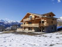 Vue de côté d'un grand chalet isolé de 3 étages en bois et pierre, grandes terrasses en bois et baies vitrées, paysage montagnard