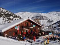 Vue du restaurant, chalet en bois, avec sa terrasse, tables et chaises, chaises lonuges. Montagnes enneigées tout autour et en arrière-plan