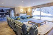 Grand salon de chalet confortable et lumineux, grand canapé d'angle, tapis, table basse, ameublement bois. Coin cuisine avec large TV et porte-fenêtre