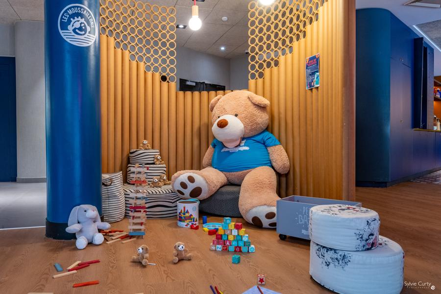 Un coin pour enfant avec une peluche géante, des kapla, des cubes, des petites peluches et des poufs.