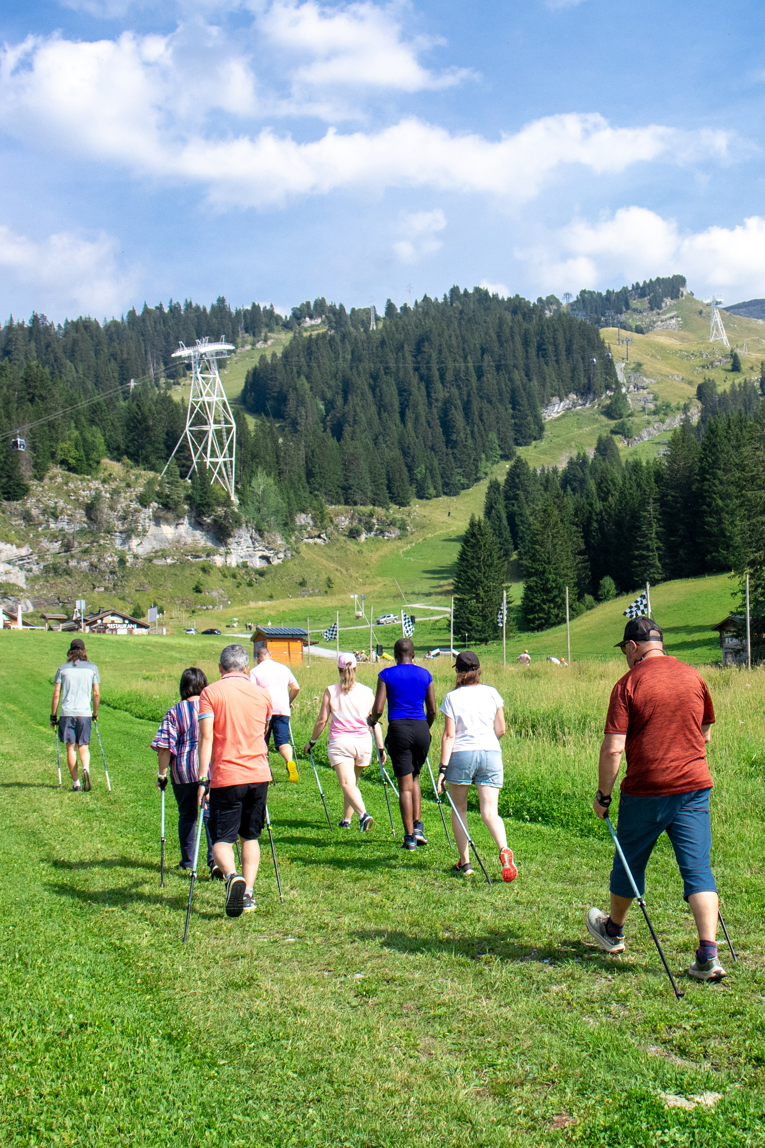Des marcheurs nordiques gravissent une pente herbeuse face à une montagne boisée couronnée d'un téléphérique.