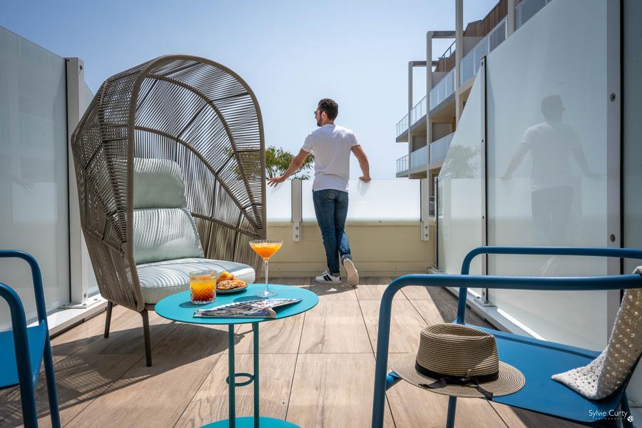 La terrasse d'une chambre avec deux chaises, un fauteuil oeuf, une table basse avec deux cocktails et cookies. Un homme regarde la vue en fond.