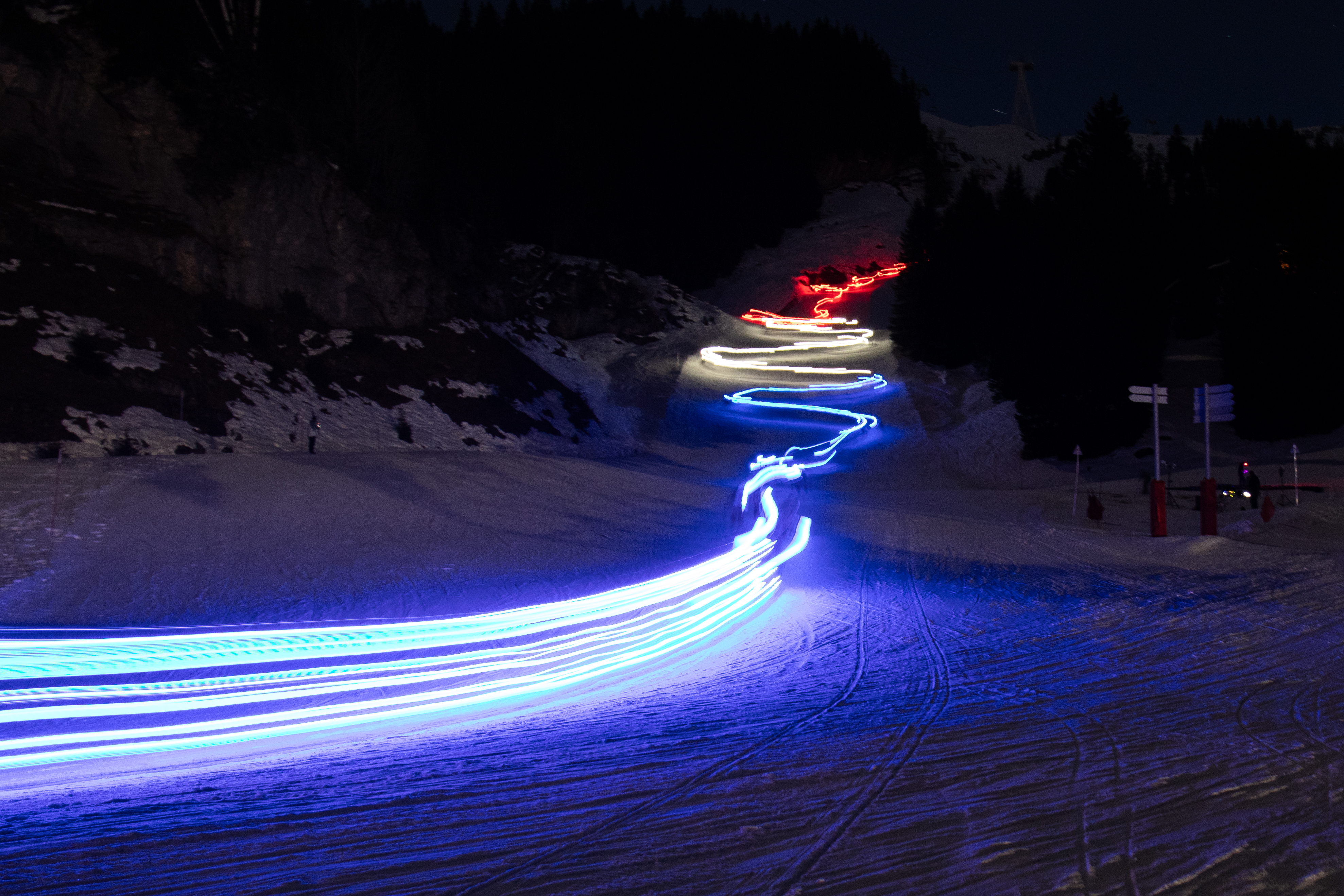 Dans la nuit de Flaine, les moniteurs de l'ESF forment un drapeau français en mouvement. Une traînée de lumières bleu, blanc et rouge serpente sur la piste sombre, créant un ruban tricolore lumineux entre les silhouettes noires des sapins. traduit