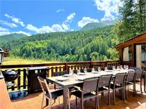 Terrasse en bois du chalet avec grande table d'extérieur, transat, véranda, vue dégagée avec paysage d'été