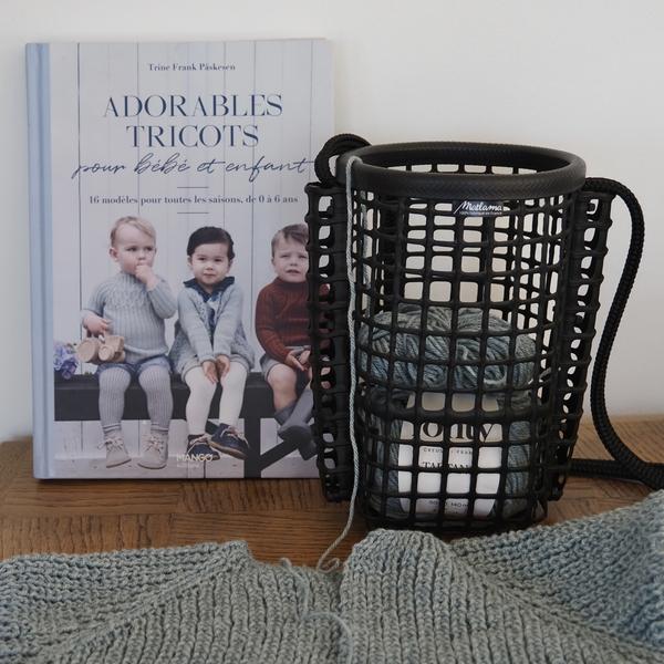 Un petit  panier en matière de poches ostéricole, avec dedans des pelotes de laine, à côté d'un livre "Adorables tricots".